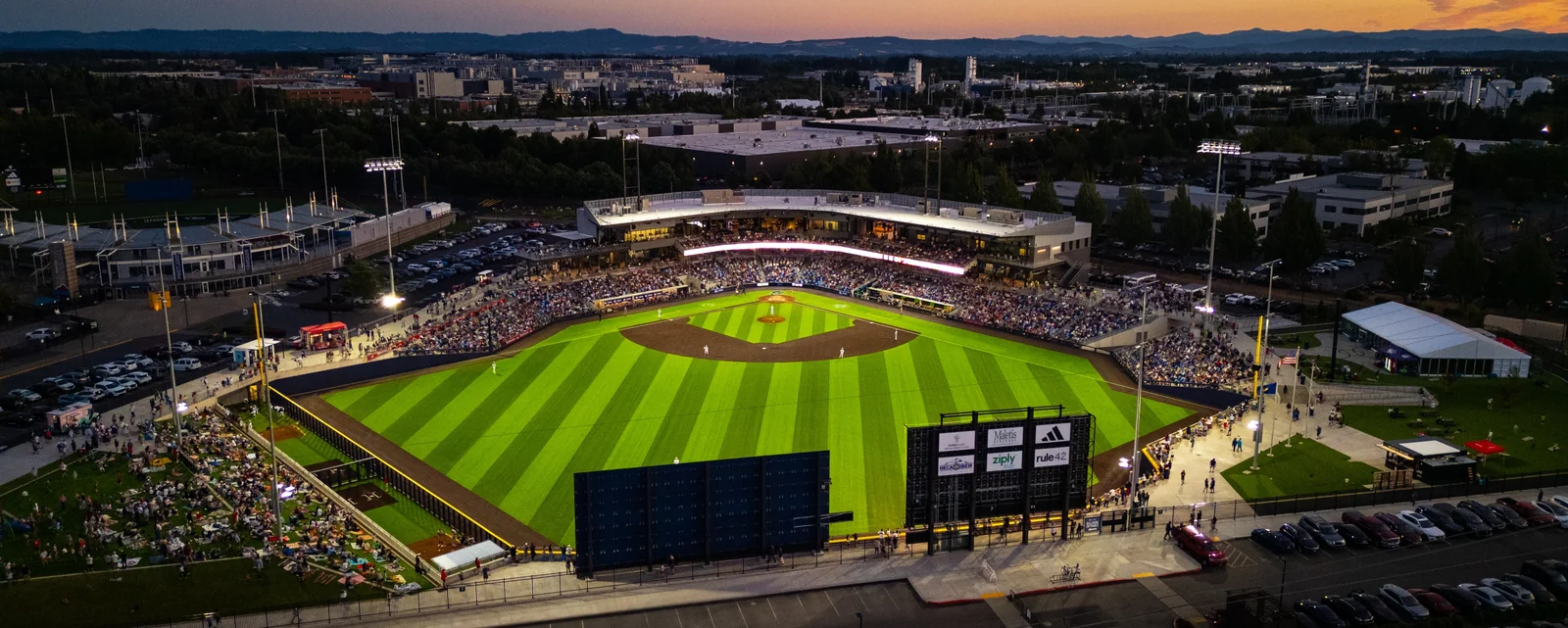 Hillsboro Hops Ballpark from the air at dusk: a full house under the lights, the striped outfield grass, the videoboard, and the lawn seating beyond the left-field wall.